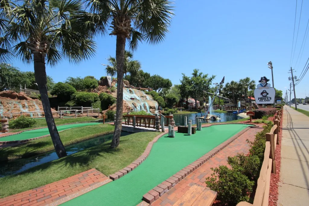 A well-manicured mini-golf green with trees and a waterfall in the background. Mini-Golf can be one of the more popular RV park amenities.