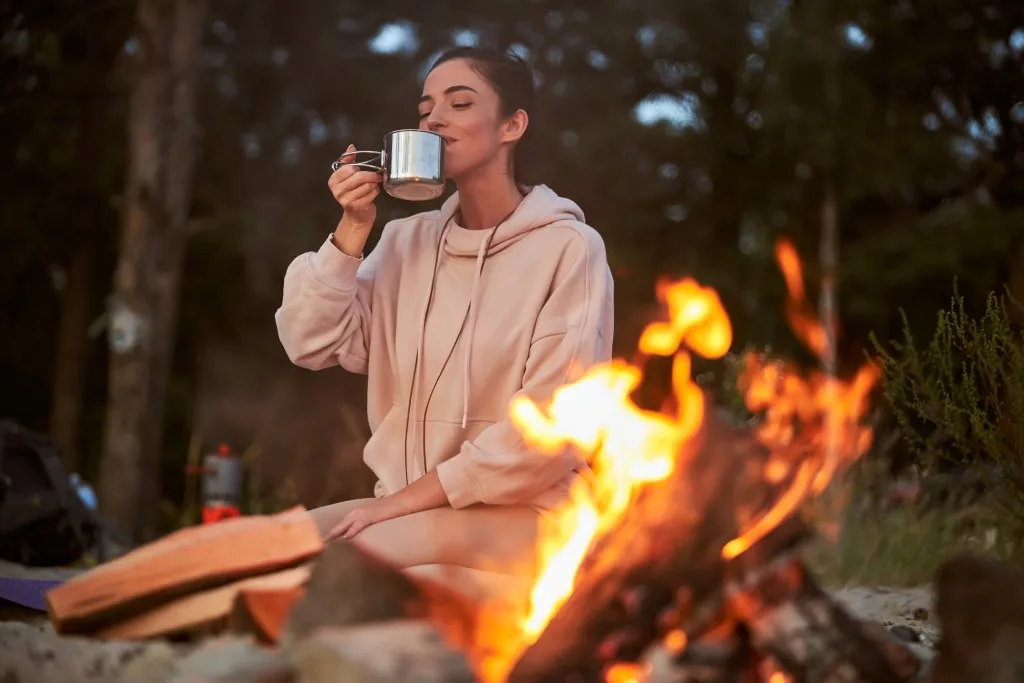Charming female traveler enjoying hot drink while resting near bonfire in the woods