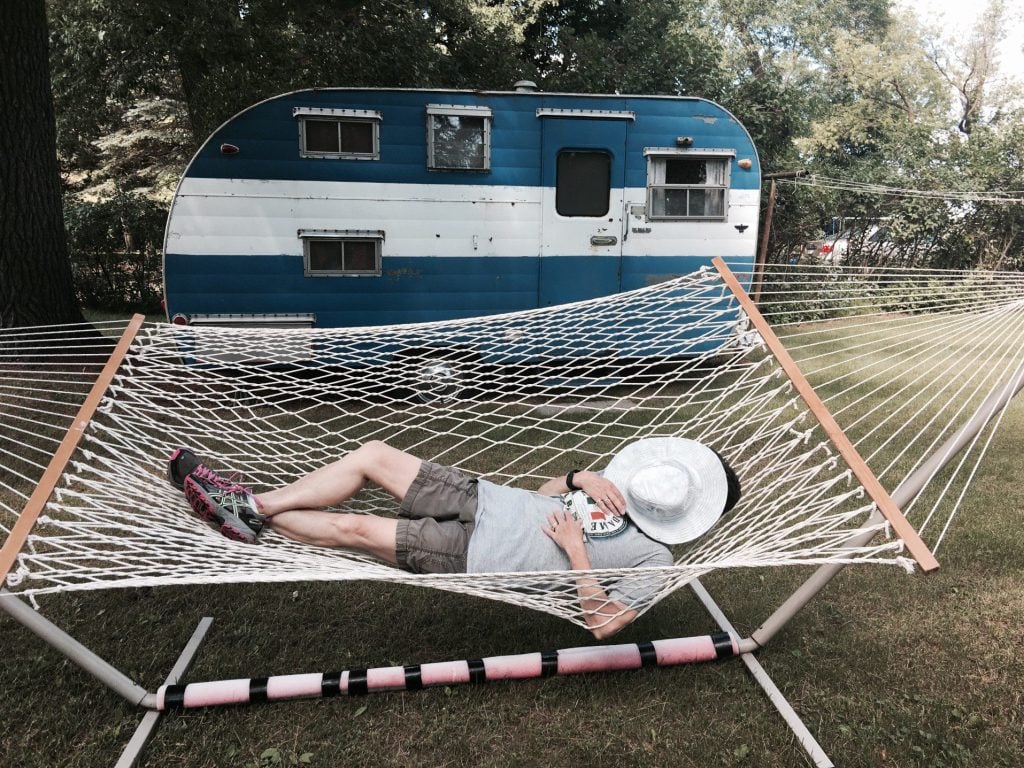 Man napping in hammock in front of vintage trailer.