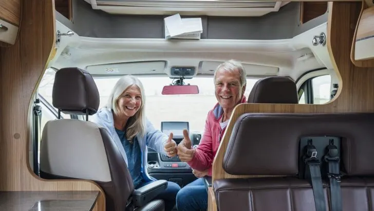 A shot of a senior couple in their camper van in Cornwall. They are both sat in the front seats looking back at camera with their thumbs up to the camera.