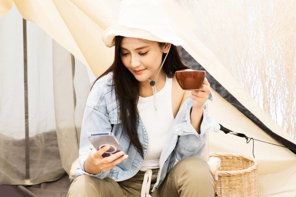 Woman looking at phone while camping and drinking coffee