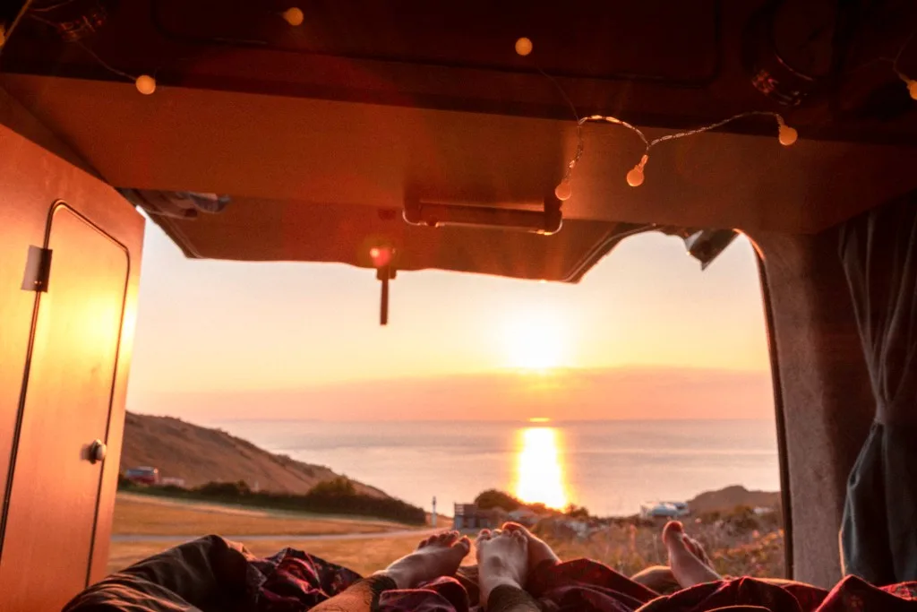 Couple stealth camping inside camper van looking at sunset over ocean