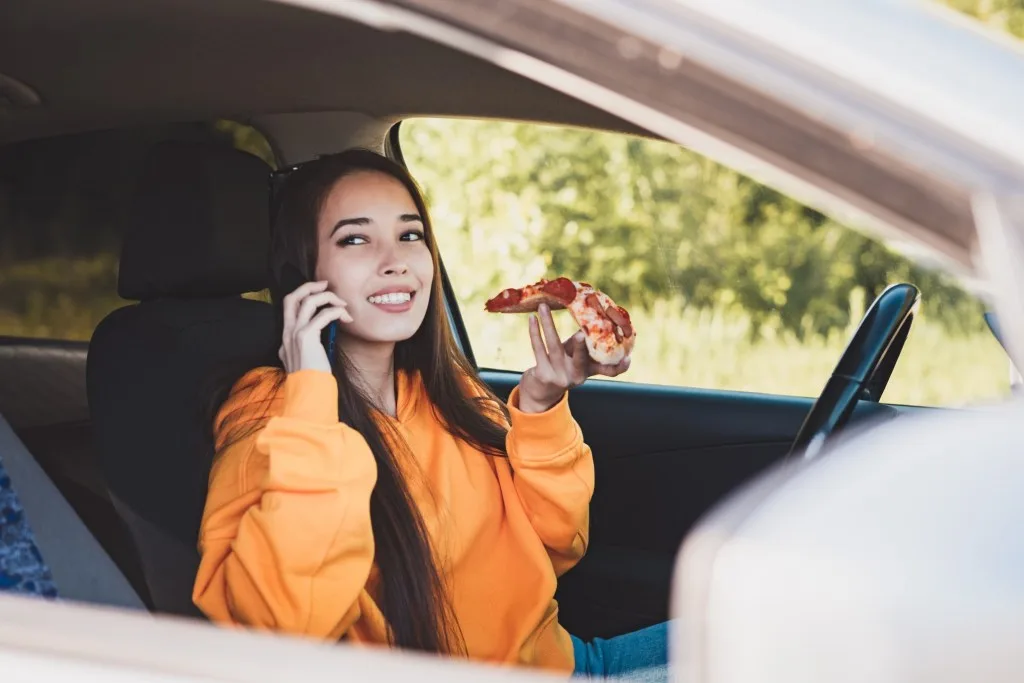 Woman eating slice of pizza in car while on road trip
