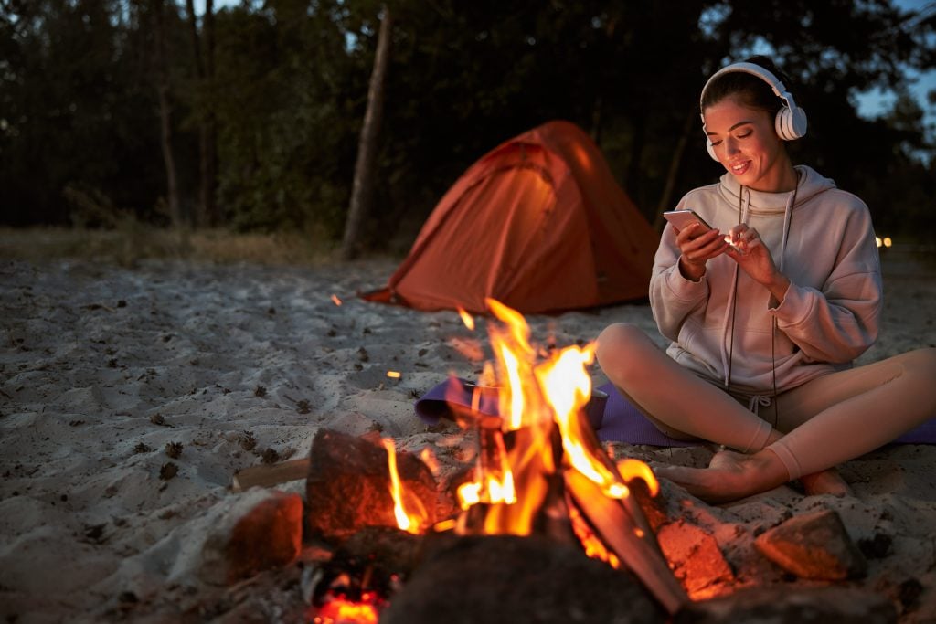 Beautiful female traveler in wireless headphones holding smartphone and smiling while resting on sand near bonfire
