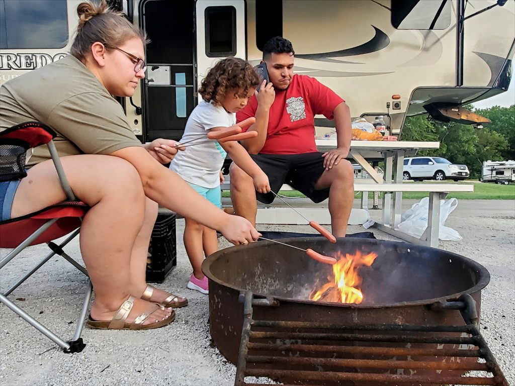 Family making hot dogs over campfire in front of RV