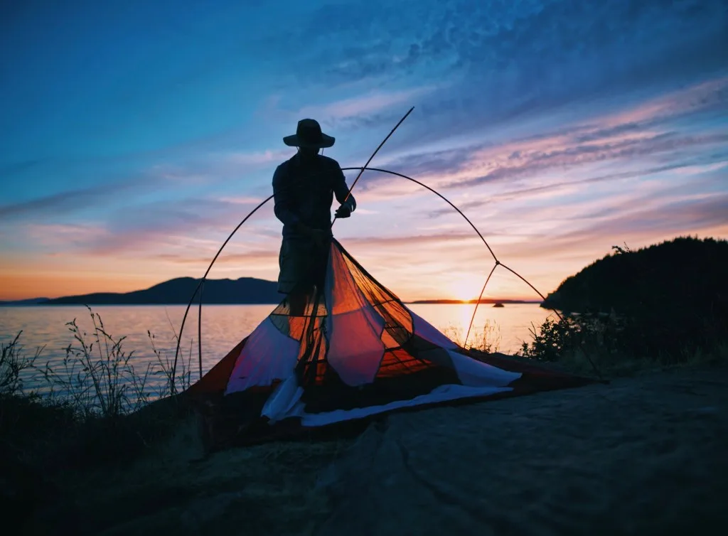 Man pitching tent next to lake at sunset