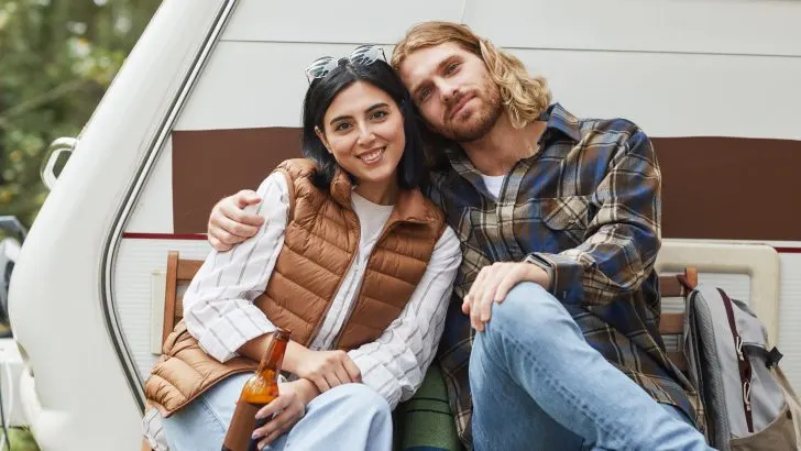 Portrait of young couple relaxing outdoors while camping with trailer van and looking at camera, copy space