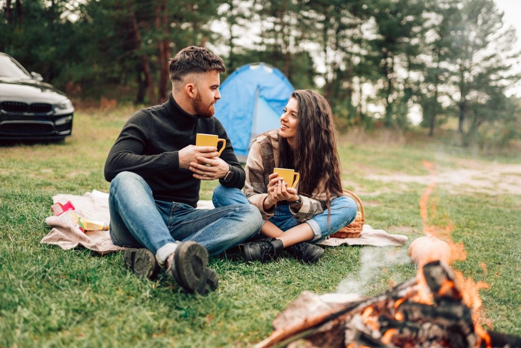 Young couple in nature sitting near campfire drinking coffee while camping