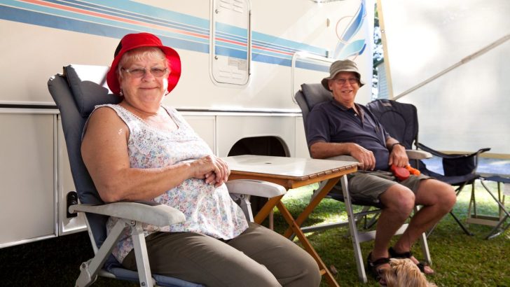 Happy retired senior couple sitting next to their motor home or RV with their dog.