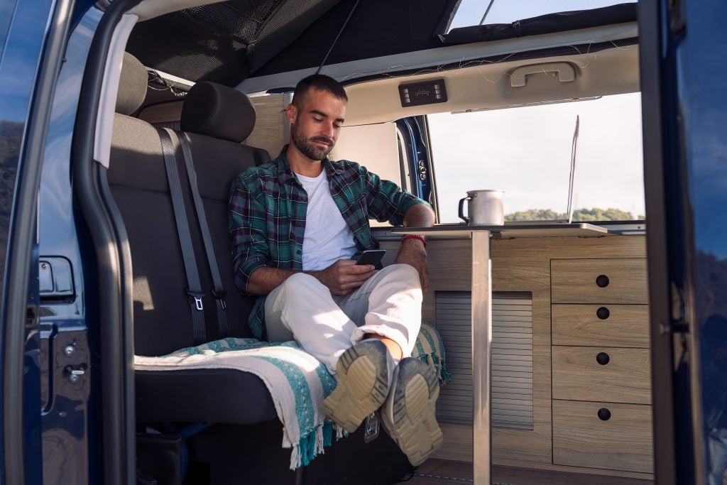 young man sitting inside his camper van using his mobile phone, concept of freedom and digital nomad lifestyle