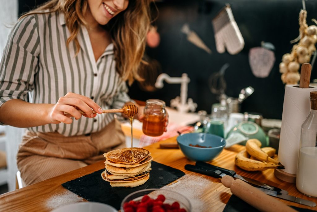 Selective shot of smiling young female food blogger and chef adding honey syrup on pancakes in modern kitchen