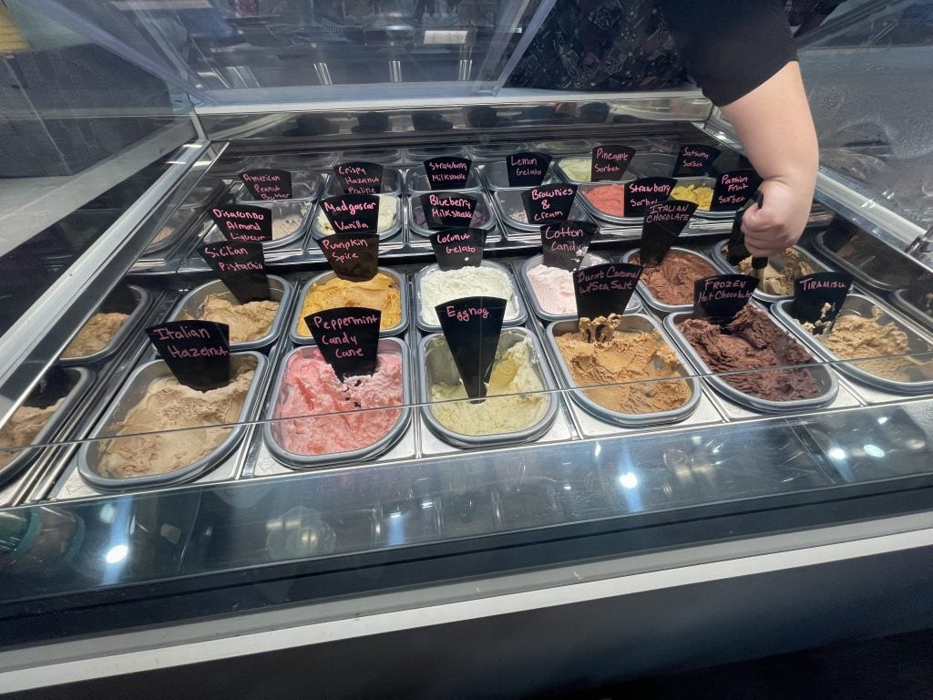 A photo of various gelato flavors in a refrigerated case. Someone is scooping the espresso flavor for a customer.