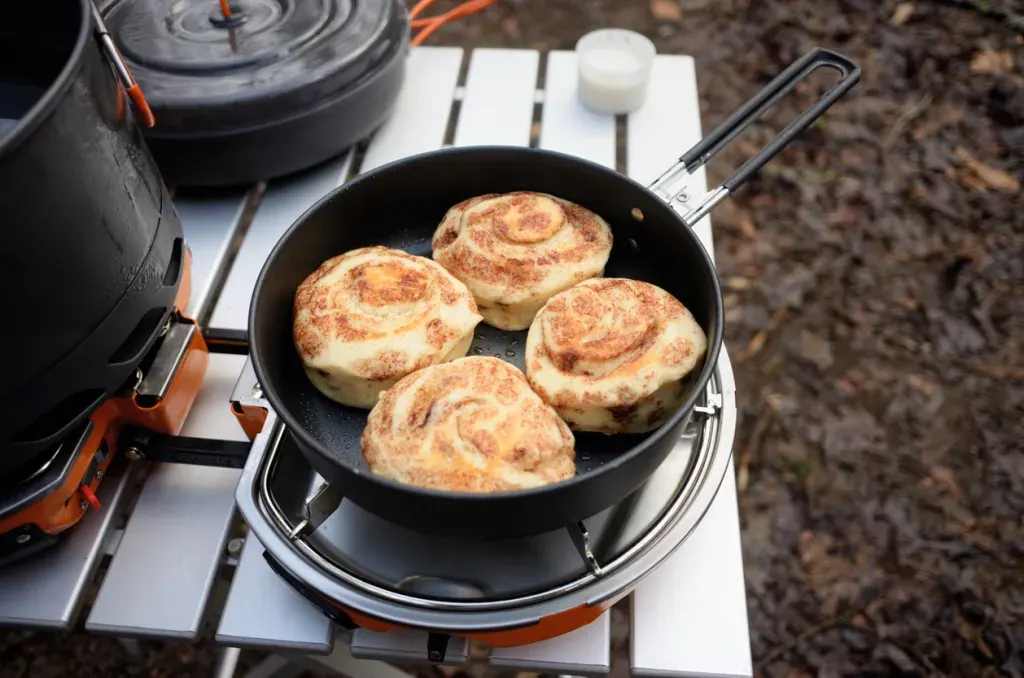 cinnamon rolls cooking in a skillet on a gas stove likely used while someone is car camping