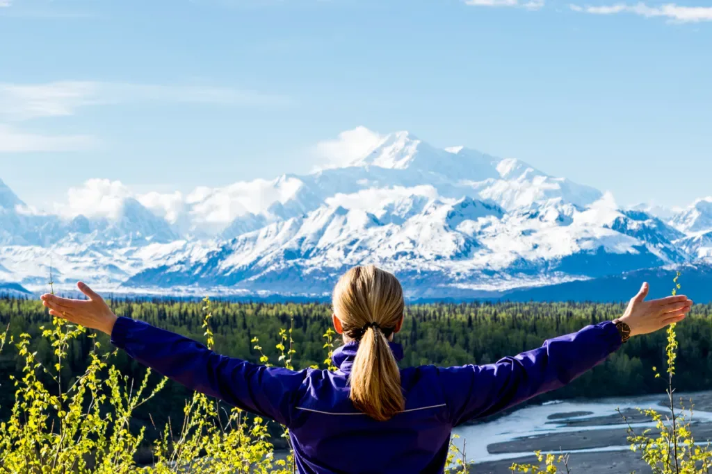 Woman with arms outstretched looking at Mount Denali on a sunny day. This may be her toughest highpointing trip.