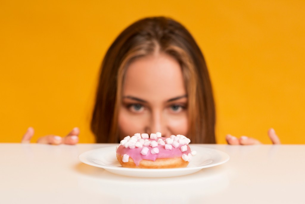 Hungry woman peeking out of behind table and looking at doughnut, one of the guilty pleasures found in Chicago