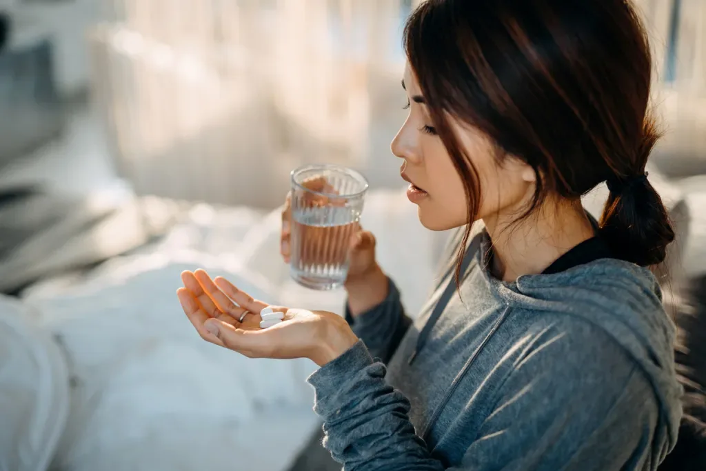 Young Asian woman sitting on a bed taking medicines in hand with a glass of water. She may be taking antibiotics to get rid of the leprosy bacteria.