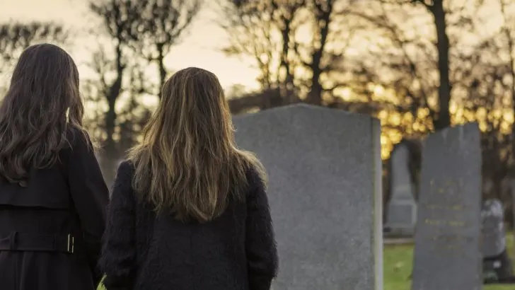 Two women with their backs to the camera look up on a tombstone. Are they visiting Bob Ross's grave?