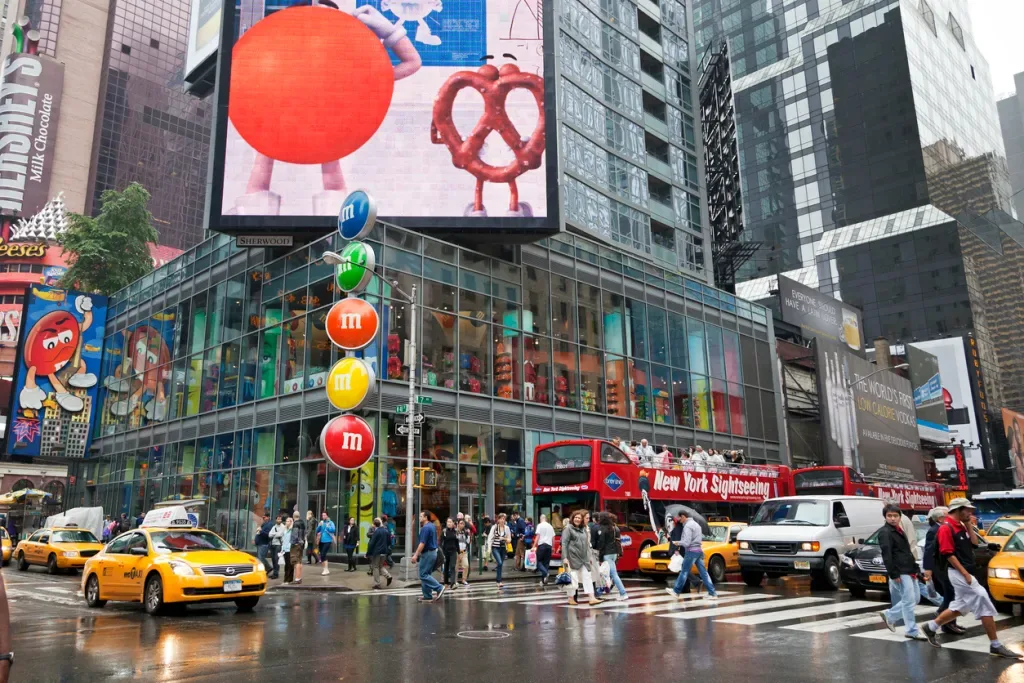 People walking on Times Square along the stores, cars and taxi's passing by and other traffic and a sightseeing bus are waiting for the traffic lights. 