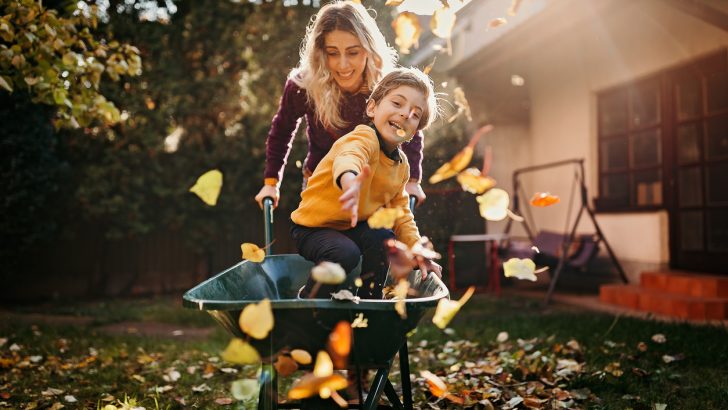 A woman giving a boy a ride in a wheelbarrow with autumn leaves falling around them