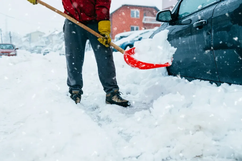 Man in red jacket clearing snow from around his car with a red plastic shovel. Think about becoming a snowbird and flying to a sunnier destination.