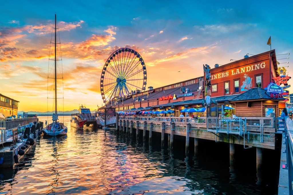People walk and dine at the downtown Seattle waterfront with the landmark Great Wheel in the background, at sunset time. Our Washington road trip begins in Seattle.