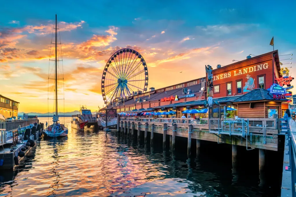People walk and dine at the downtown Seattle waterfront with the landmark Great Wheel in the background, at sunset time. Our Washington road trip begins in Seattle.