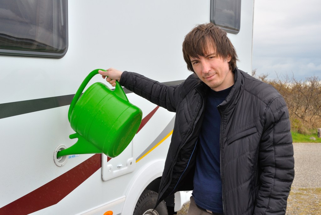 A man filling his water tank using a garden watering can. You'll need to flush your tanks when you're ready to camp again if you pour antifreeze down your RV drains.