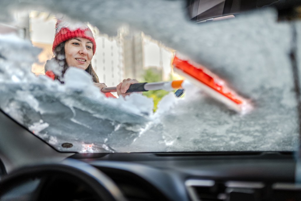 Millennial Woman in winter jacket scraping ice and snow from car windows before driving