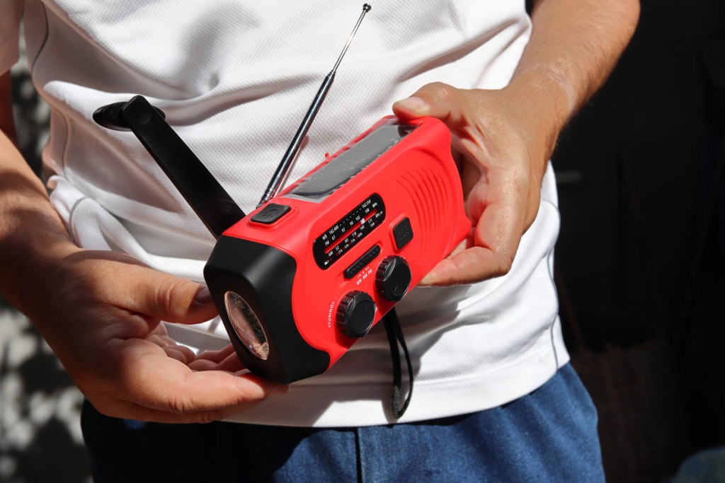 A man holds in his hands a small red portable emergency radio rechargeable with solar panels or manually with a crank and an included flashlight