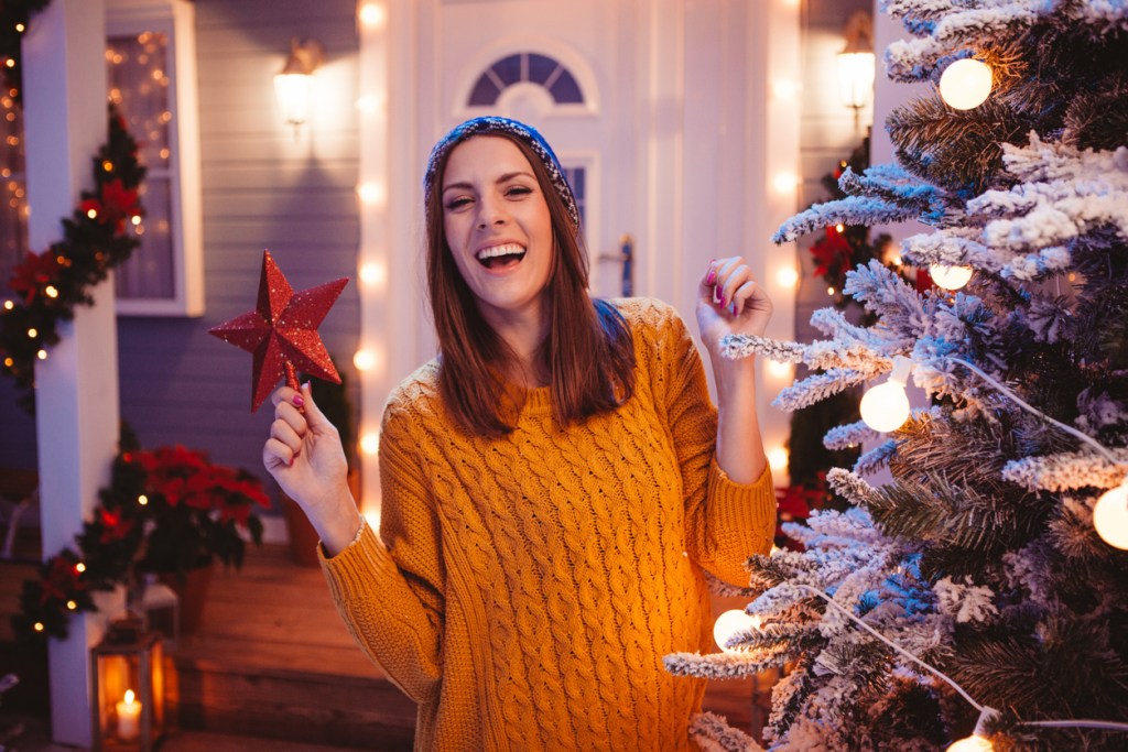 Young woman in front of house, trying to reach the top of a tree to put star on it. Wearing knitted sweater and hat. House, yard and tree are decorated with festive string lights. Evening or night with beautiful yellow lights lightning the scenes.