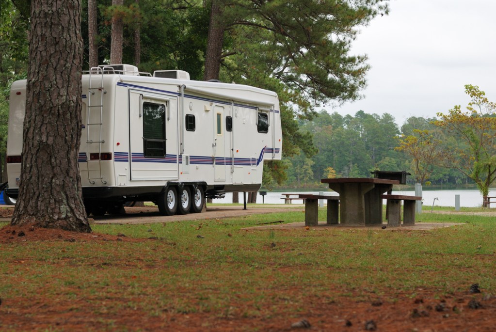 A 5th wheel camper parked under trees by a lake - Are you ready for a new RV in 2024?