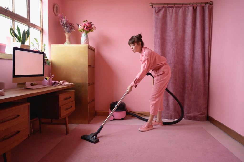 A woman dressed in pink vacuuming a pink carpet with a pink vacuum in a pink room - don't make the mistake of not cleaning your vacuum