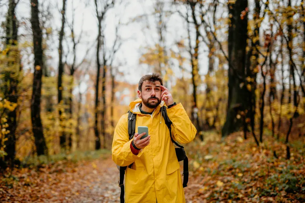 A man in yellow jacket hiking in the woods, carrying a cell phone and looking lost