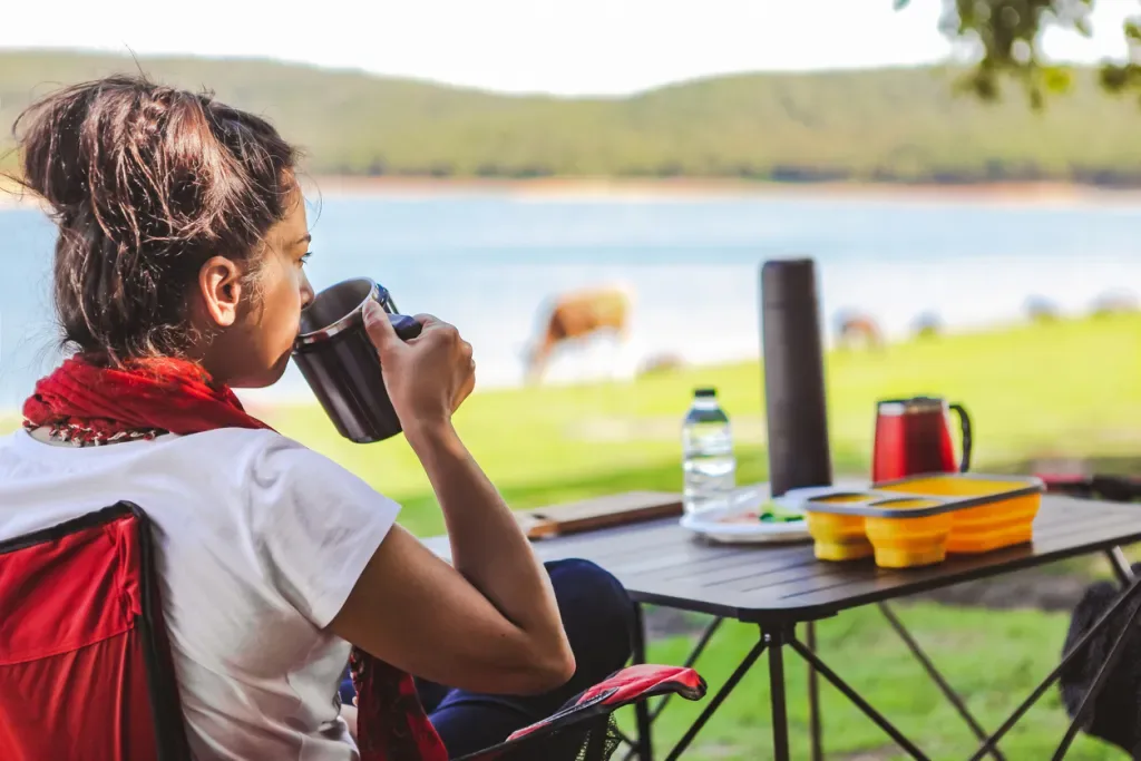 A woman enjoys her coffee at a camping table by a lake.