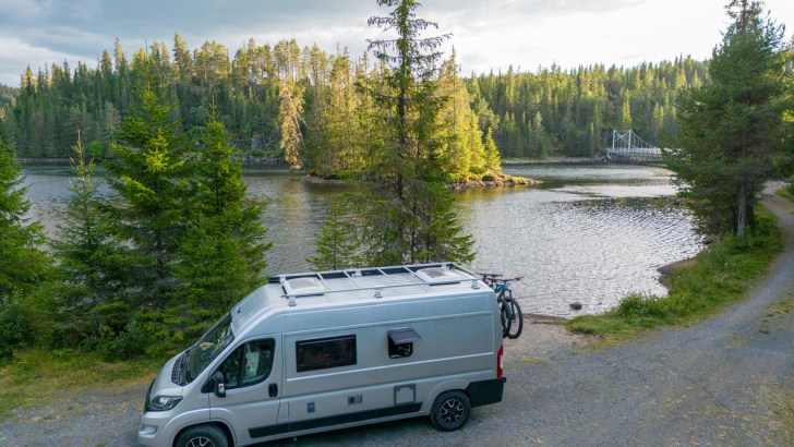 A silver off-grid camper van parks by a serene lake.