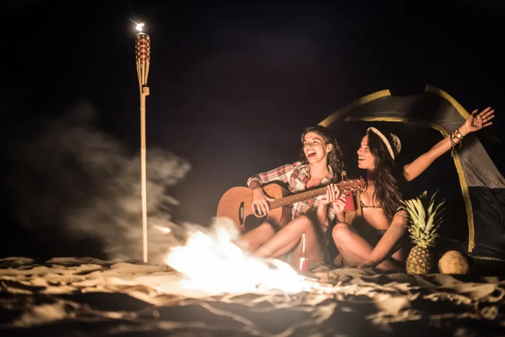 Two women play music on the beach by a fire. An iconic TIKI Torch burns nearby.