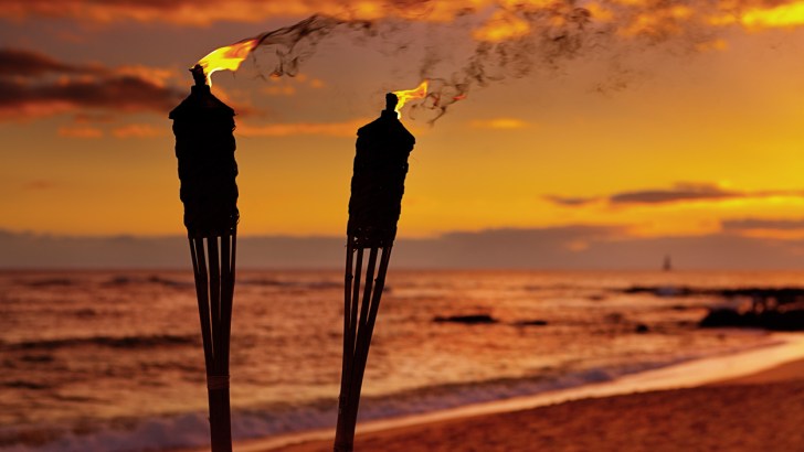 Two TIKI Torches, the brand's most iconic product, burning on the beach by the ocean at sunset.