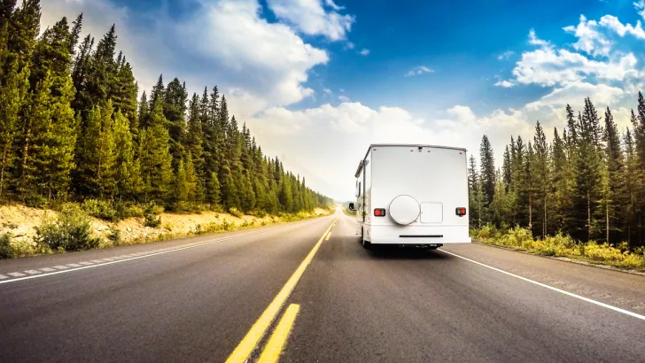 An RV barrels down an open road flanked by evergreen trees on a beautiful day.