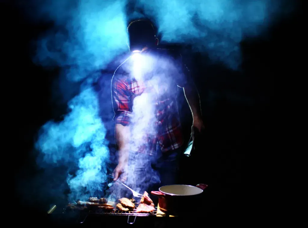 A man wearing a headlamp stand over a grill. A Magna Grill Light would make this experience much more enjoyable.