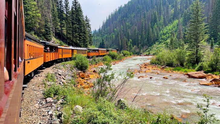 An orange train travels along the Animas River on the Durango & Silverton Narrow Gauge Railroad.