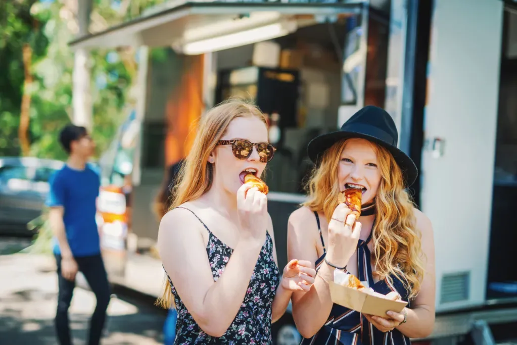 Two young women eat tasty treats at an outdoor food festival.