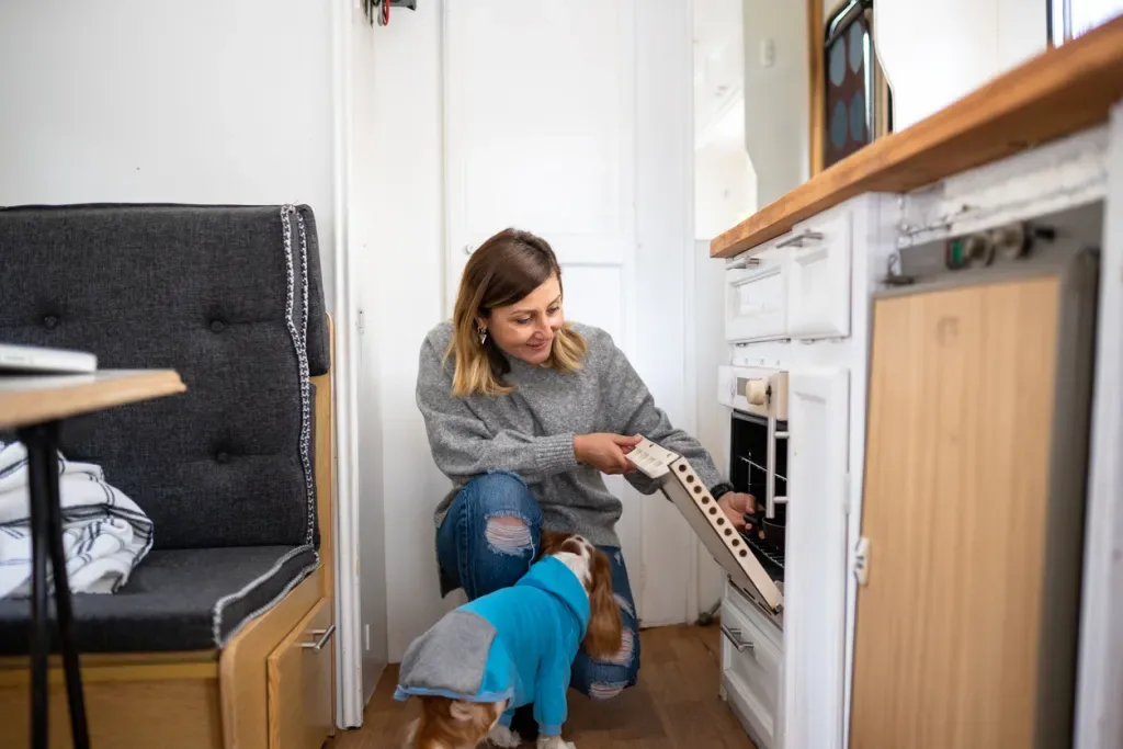 A woman using the oven in her RV with a curious dog nearby. Her clutter-free space makes it appear that she's embraced tiny living.