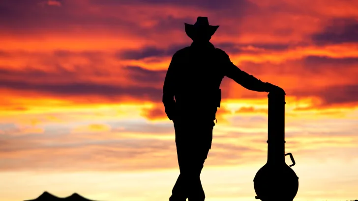 Silhouette of a man wearing a cowboy hat standing by a guitar case in front of a colorful sunset.